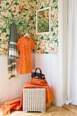 Corner of bedroom with green floral wallpaper and wicker stool against wood-panelled dado; orange dress and fringed blanket complement the colour scheme