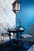 Moroccan-style pendant lamp above antique table and two wire chairs in corner of room with one blue wall and patterned wallpaper