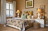 Double bed with beige, quilted headboard flanked by antique bedside lamps on white bedside cabinets