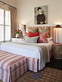 Double bed and matching ottoman in pink and white stripes with various scatter cushions on bed and portrait of woman on wall above