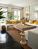 Large ottoman surrounded by white sofas with many yellow scatter cushions in bright living room with dark, glossy floor