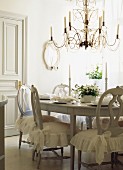 White dining room with festively set table, chandelier & chairs with romantic, flounced seat cushions