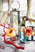 Red-painted branch in front of roses in colourful glasses and lanterns on table