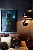 Dramatic lighting in corner of bedroom - various pendant lamps above ethnic scatter cushions on bed against dark-painted wall and photo of very dark-skinned person