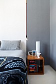 Cubic bedside cabinet made from exotic wood next to bed with pale grey bed linen against white wall; simple light bulb as pendant lamp