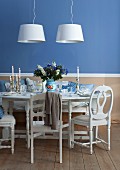 Festively set white dining table with silver candlesticks against blue wall and two modern pendant lamps