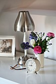 Shabby chic arrangement on bedside table with metal lamp, alarm clock and posy of spring flowers