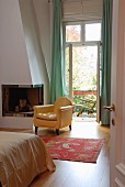 Yellow-covered, antique armchair next to fireplace in bedroom and open balcony door with a view