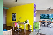 Desk and office chair against bright yellow wall in open-plan, colourful living area with dining table in background