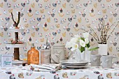 Napkins, tablecloth and wallpaper with colourful hen pattern and grey crockery with hen-motif relief on table set for Easter