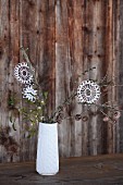 White crocheted stars hanging from twigs in white china vase in front of rustic wooden wall; vintage Christmas atmosphere