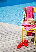 Strohhut und pinkfarbenes Handtuch auf gelbem Stuhl, davor rote Damen-Sandalen auf sonnenbeschienener Holzterrasse am Pool