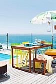 Wooden table, matching bench and yellow chairs on sunny wooden terrace beside pool with sea view through glass balustrade