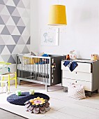 Pale grey chest of drawers and cot against wall with white and grey geometric pattern; yellow pendant lamp above rug and cushion on floor