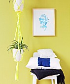 Green plant in yellow macrame plant hanger next to stack of cushions on chair against lime green wall