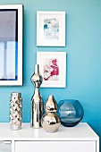 Verspiegelte Vasen in verschiedenen Grössen auf weißem Sideboard vor hellblau getönter Wand mit gerahmten Bildern
