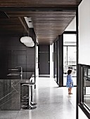 Long corridor in minimalist house with dark grey wall elements and window frames, stone-clad island counter and bar stools in kitchen