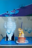 Retro serving trolley with white Michelin Man figurine, donuts and glasses of sparkling wine in front of modern, pale blue pixellated picture