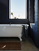 White vintage bathtub below window and grey-painted brick walls
