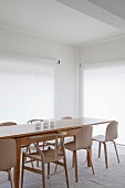 Long, Scandinavian-style, designer wooden table and wooden chairs on pale rug and large windows with white, translucent roller blinds