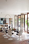 White tulip chairs with dark seat cushions around oval glass table below arc lamp and open lattice French windows to one side in modern interior