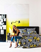 Blonde woman perched on bed with grey, floral bed linen, sconce lamp mounted on wall with white and yellow ombr