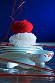 Asiatische Tischdeko: Schälchen zwischen gespanntem, rotem Wollfaden, im Hintergrund Glasnudeln und roter Wollknäuel auf Tellerstapel