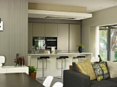 Dining area next to sofa with scatter cushions in front of open-plan fitted kitchen with designer bar stools at breakfast bar
