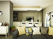 View across grey sofa with scatter cushions to open-plan kitchen area with white, fitted kitchen and bar stools at counter
