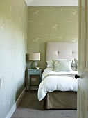 View through open door of bed with upholstered headboard and table lamp on small bedside table against wallpaper with pattern of birds