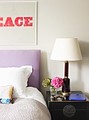 Vintage lamp on bedside cabinet next to double bed with lilac headboard and text artwork on wall