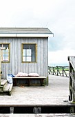 Cushions on bench and beach bag on wooden platform in front of grey, weathered wooden beach hut
