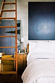 Bett mit weißer Betwäsche und dunklem Bild am Kopfende, lange angelehnte Holzleiter und alte Holzkiste als Nachtkästchen