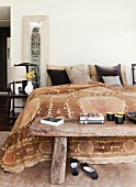 Rustikale, chinesische Holzbank am Bettende eines schlichten Doppelbettes mit folkloristischer Tagesdecke in Brauntönen und Kissen