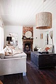 Stacks of scatter cushions on ecru sofa, dark wooden trunk on wooden floor, pendant lamp with large cylindrical lampshade hanging from white wooden ceiling and rustic fireplace