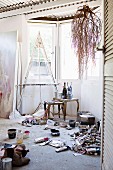 Corner of studio - used tubes of paint and painting utensils on floor, easel and side table in window bay, branches hung from wooden beams below corrugated metal ceiling