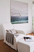 Comfortable white loose-covered armchair, reading lamp, pouffe and peaceful seascape painting