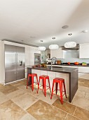 Designerküche mit Küchenblock, roten Barhockern und drei Designerpendelleuchten, Natursteinfliesenboden und Edelstahlschranktüren