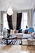 Illuminated ceiling lamp with white lampshade above round coffee table, corner sofa with many scatter cushions and rug with ethnic pattern on wooden floor
