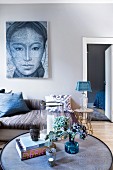 View past vase of flowers on round coffee table to couch below large portrait on wall painted pale grey