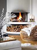 Cosy basketwork armchair with white fur blanket in front of fire in open fireplace with firewood store below