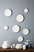 Stacked, simple china crockery and patterned, lidded urn below white plates on grey wall