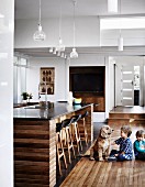 Two children playing with dog on wooden floor next to island counter below pendant lamps in open-plan kitchen