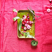 Red and pink buttons in metal dish with tape measure and scissors