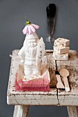 Still-life arrangement of white, china garden gnome wearing flower hat, feather with lettering and cutlery on stool with peeling paint