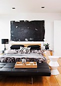 Black leather couch at foot of double bed and white stool on wooden floor in modern bedroom with black modern artwork on wall