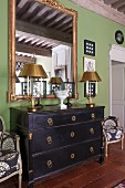 Table lamps with gold lampshades on black-stained chest of drawers below mirror on green wall