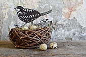 Hand-crafted paper bird and quail eggs in birds' nest on wooden surface against wall with peeling paint