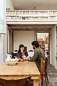 Young couple sitting at rustic wooden table on old, grey stone floor; view into workshop and of wooden mezzanine with bookcase balustrade