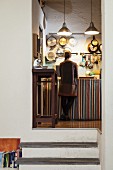 Blick in erhöhte Vintageküche mit an der Wand hängenden Pfannen, davor junge Frau an Küchenunterschrank mit Streifenvorhang